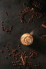 Glass of spicy pumpkin latte with whipped cream and cinnamon standing on black serving board. Coffee beans and spices above. Dark background. Top view, space.