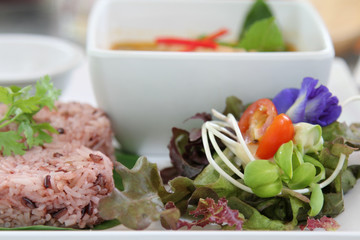 Red steamed rice on banana leaf with coriander on topped and salad with pea flower on white plate.