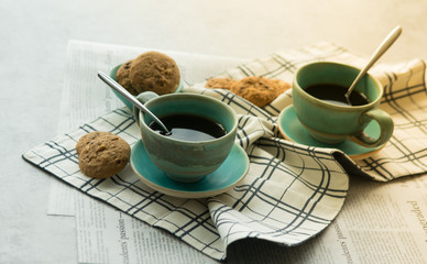 Two cups of black coffee with spoon on supported dish on fabric on black and white newspaper are put on hard table in the early morning with bright yellow light and some biscuits.