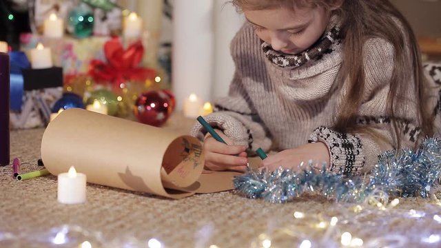 cute little girl lies on the floor near the Christmas tree and draws a greeting card