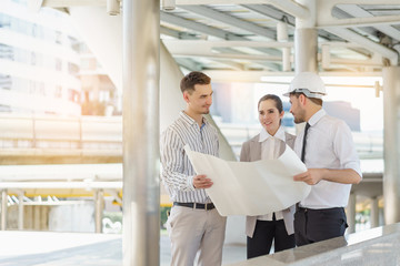 Three people are business manager and engineer meeting project at construction site. Man wearing safety helmet and holding blueprint document.
