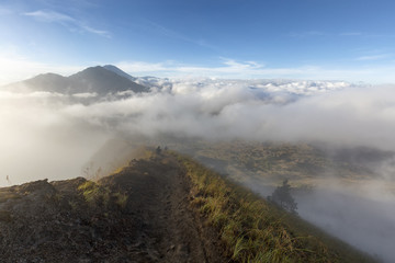 A trail leads into the fog at the rim of the caldera on Mount Batur with Mount Agung in the Background in Bali, Indonesia.