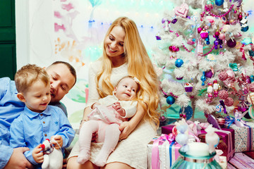 Obraz na płótnie Canvas Beautiful family with children in warm sweaters poses before a wall and rich Christmas tree