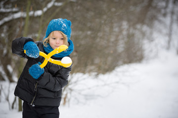Portrait of adorable little kid boy with long blond hair playing with snowballs outdoors..Child with blue scarf and hat walking and having fun on a windy winter day.