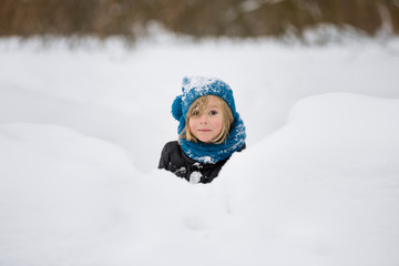 Fototapeta na wymiar Portrait of adorable little kid boy with long blond hair playing with snow outdoors in the park. Child with blue scarf and hat walking and having fun on a windy winter day.