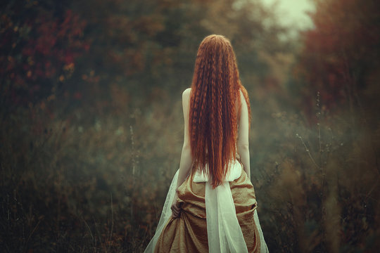 A beautiful young woman with very long red hair as a witch walks through the autumn forest. Back view.