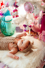 Baby in deer suit lies on soft white pillow under white Christmas tree with pink toys