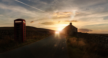 Car travelling on rural road with telephone box and house at sunset
