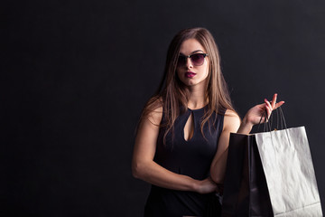 Fototapeta na wymiar Elegant brunette woman wears sunglasses and black dress holding black shopping bags, black friday concept