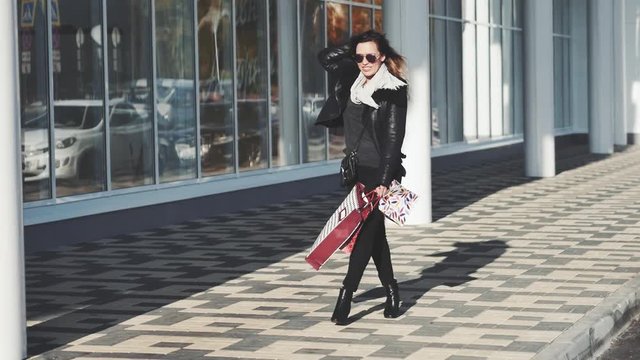Beautiful girl in sun glasses a black leather jacket, black jeans is holding shopping bags, looking at camera and smiling while walking down the street