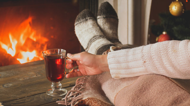 Toned closeup image of young woman holding cup of tea at burning fireplace in living room