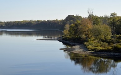 curvy pebble shoreline at a lake lined with with colorful trees in Autumn reflecting on the water surface with a single boat on a dock