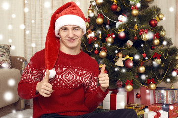 Young man dressed in retro sweater and sanat hat sit near christmas tree. New year eve, ornated fir tree. Winter holiday and love concept.