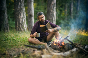 Hipster hiker with book and mug at bonfire in forest