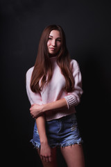 Model test of beautiful brunette woman with long hair, wears pink sweater,short blue shorts, posing on a black background