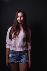 Model test of attractive brunette model with long hair, wears pink sweater,short jeans shorts, posing on a black background