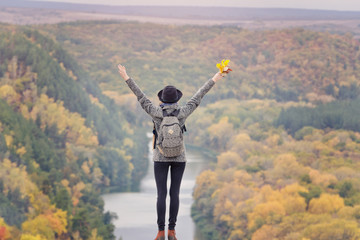 Girl with a backpack and a hat standing on a hill. Hands raised up. River and mountains below. Back view