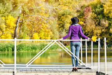 Girl in hat standing on the bridge near the water. Sunny autumn. Back view