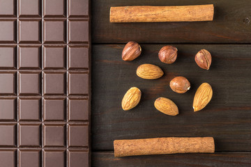 Bitter, dark chocolate bar, almond, hazelnut and cinnamon stick on wooden background, top view