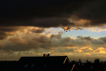 Samolot awionetka w burzowych chmurach nad dachami budynków. - obrazy, fototapety, plakaty