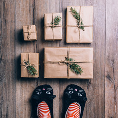 Feet with warm winter socks and cat slippers standing in front of Christmas gifts. top view.