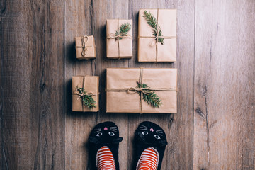 Feet with warm winter socks and cat slippers standing in front of Christmas gifts. top view.