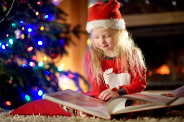 Fototapeta na wymiar Happy little girl reading a story book by a fireplace in a cozy dark living room on Christmas eve