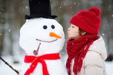 Adorable little girl building a snowman in beautiful winter park. Cute child playing in a snow.