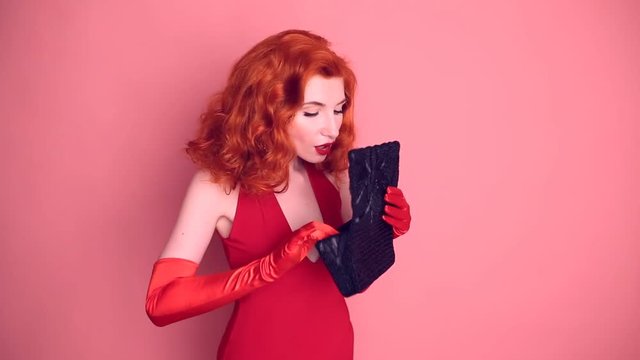 Luxurious red-haired retro woman with expensive jewelry in a red dress and fur cape posing on a pink background. A smart girl in long silk gloves looks into a black open bag