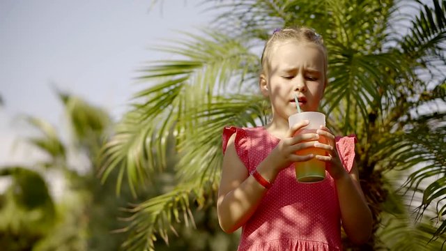 A small and funny girl drinks fruit juice from a plastic cup with a straw, the child enjoys a soft drink