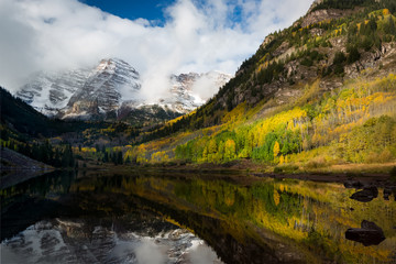 Fototapeta na wymiar Maroon Peak and its reflection on Maroon Lake and aspen trees with its gold yellow leaves in fall foliage autumn season in a bright day light sunny day cloudy blue sky, Aspen, Colorado, USA.