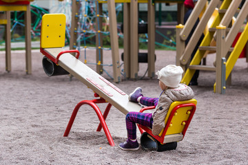 Little cute caucasian girl in jacket and hat sitting alone on a seesaw swing at a playground...