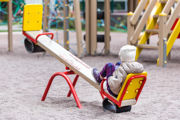 Little cute caucasian girl in jacket and hat sitting alone on a seesaw swing at a playground...