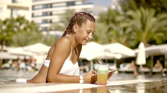 An adult teenager holds a portable tablet in his hand, a glass of juice is standing next to him, a lady is resting in the pool