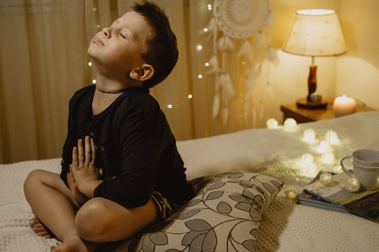 Cute boy praying for gift with eyes closed in the evening room on a bed on a background of lights