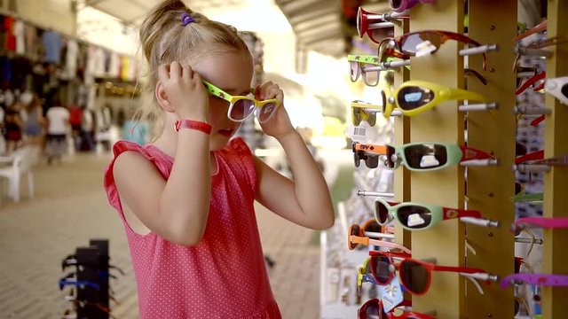 funny small girl is trying on childrens sunglasses in a shopping mall, she is likes it, smiling