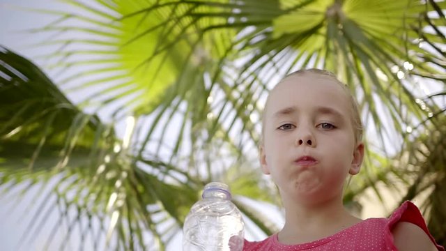 little caucasian girl is drinking pure water from bottle in summer heat, standing under big palm