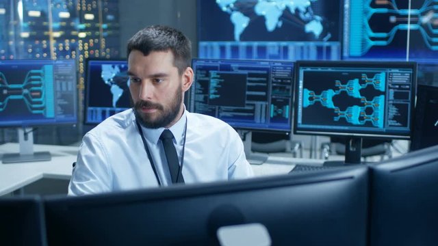 In the System Control Center Technical Operator Monitors Displays at His Workstation. He's Surrounded by Displays Showing Relevant Data. In the Background Data with Interactive Map.