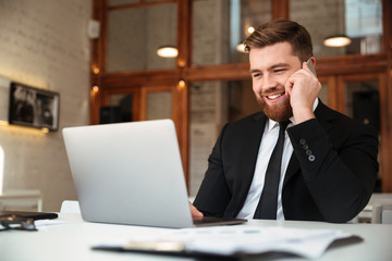 Happy young businessman in black suit talking on mobile phone, looking at laptop while sitting at his workplace