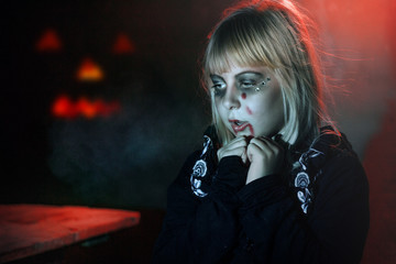 little girl with blond hair in the make-up for Halloween is standing in a dark room near the pumpkin