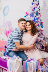 Cheerful couple sits hugging before a Christmas tree in pink room