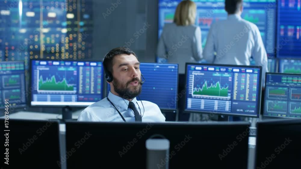Aufkleber Stock Exchange Trader Makes a Deal with Big Client Over the Headset. He Works with Other Brokers and is Surround By Computers with Graphs and Ticker Numbers on Screens. 