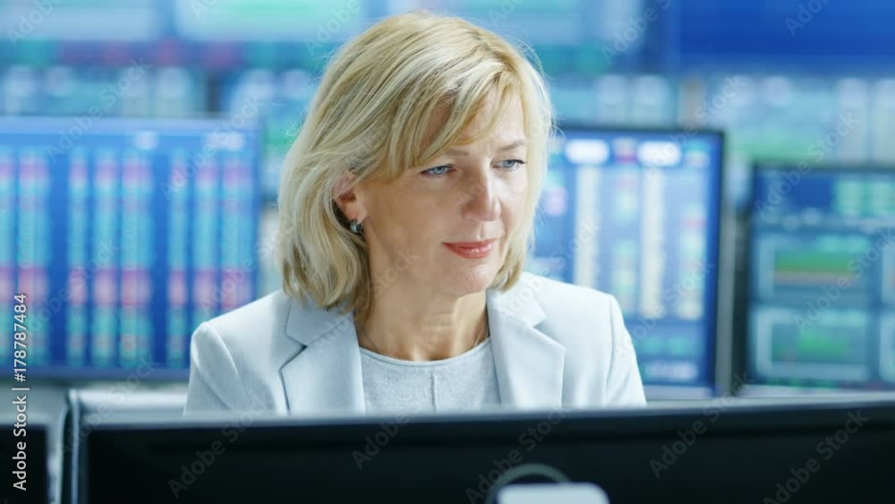 Poster Portrait of the Senior Female Stock Trader Operating at Her Workstation. Behind Her Multiple Monitors Showing Data, Ticker Numbers and Graphs. Shot on RED EPIC-W 8K Helium Cinema Camera.