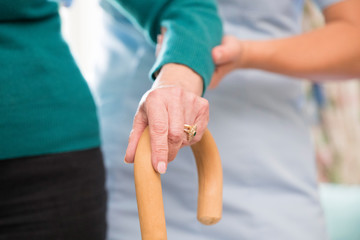 Senior Woman's Hands On Walking Stick With Care Worker In Background