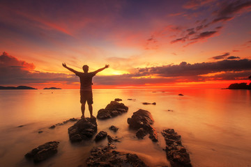silhouette of happy man standing on rock in the sea with sunset sky, long speed exposure,Tarn-khu beach at Trat, Thailand - obrazy, fototapety, plakaty