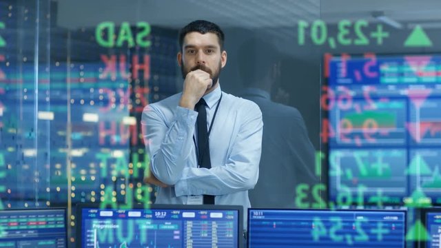 Stock Market Top Trader Looks at Projected Ticker Numbers and Graphs Running, Analysing Data to Make Best Sell. Behind Him Room Full of Screens and Statistics. 