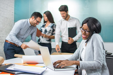 Smiling young business woman working on laptop with busy colleagues in the background.