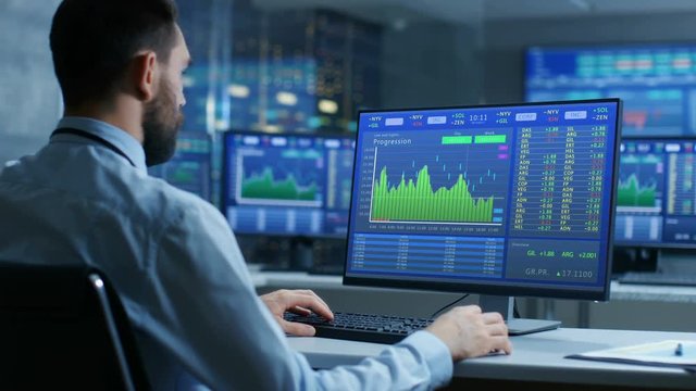 Over the Shoulder View of Stock Market Trader Working on a Computer Showing Stock Ticker Numbers and Graphs.  Shot on RED EPIC-W 8K Helium Cinema Camera.