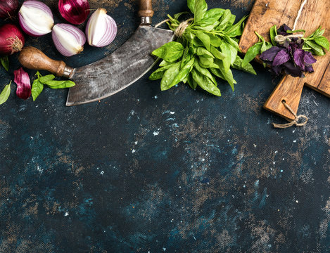Healthy food cooking background. Fresh green and purple basil leaves, red onions and garlic, herb chopper knife and rustic cutting board over grunge dark blue plywood texture. Top view, copy space