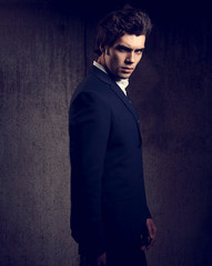 Fototapeta premium Sexy handsome male model posing in blue fashion suit and white style shirt looking down on dark shadow background. Toned portrait
